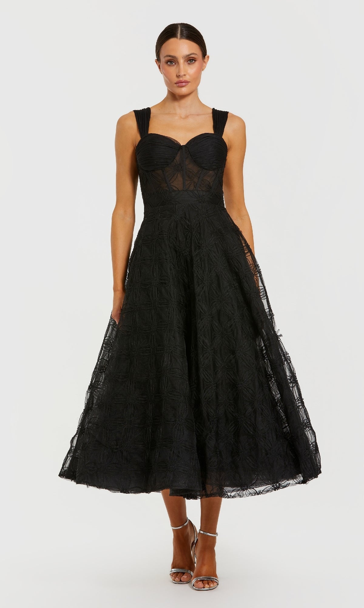 A woman wears the Mac Duggal 21171 tea-length black formal dress, featuring a fitted bodice and flared skirt, paired with strappy heels, set against a plain background.
